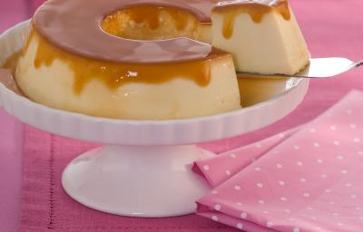Foto de uma bancada rosa, com um tecido rosa de bolinhas brancas. Ao centro há um prato branco alto com um Pudim com calda de caramelo e um pedaço dele está sendo retirado com uma espátula. Ao fundo há 3 pratos brancos empilhados. Foto de uma bancada rosa, com um tecido rosa de bolinhas brancas. Ao centro há um prato branco alto com um Pudim com calda de caramelo e um pedaço dele está sendo retirado com uma espátula. Ao fundo há 3 pratos brancos empilhados.