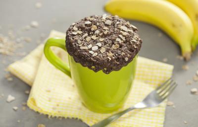 fotografia em tons de cinza, verde e marrom de uma bancada cinza vista de cima. Contém um pano amarelo, uma xicara verde com um bolo de chocolate com cereais por cima e um garfo ao lado e um cacho de bananas