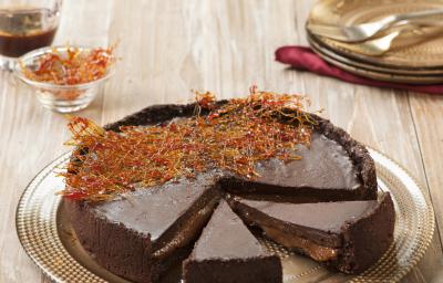 Fotografia em tons de marrom e chocolate, mostrando bancada em tons de marrom e branco contendo prato dourado com uma torta de chocolate decorada com fios de caramelo, ao fundo pratos e garfos, potinho com calda de caramelo e potinho com fios de caramelo. Fotografia em tons de marrom e chocolate, mostrando bancada em tons de marrom e branco contendo prato dourado com uma torta de chocolate decorada com fios de caramelo, ao fundo pratos e garfos, potinho com calda de caramelo e potinho com fios de caramelo.