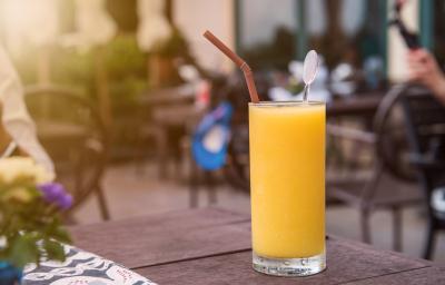 Fotografia de um copo grande com um refresco de laranja, dentro do copo, um canudo de cor marrom e um enfeito de vidro. O copo está sobre uma mesa de madeira na cor marrom. Ao fundo, mesas e cadeiras de uma cafeteria ao ar livre. Fotografia de um copo grande com um refresco de laranja, dentro do copo, um canudo de cor marrom e um enfeito de vidro. O copo está sobre uma mesa de madeira na cor marrom. Ao fundo, mesas e cadeiras de uma cafeteria ao ar livre.