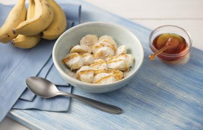 Fotografia em tons de azul em uma mesa de madeira clara com um paninho azul ao lado, um prato branco redondo fundo com as rodelas de banana cobertas com mel, aveia, leite em pó e linhaça. Ao lado, um cacho de bananas e um potinho com mel. Fotografia em tons de azul em uma mesa de madeira clara com um paninho azul ao lado, um prato branco redondo fundo com as rodelas de banana cobertas com mel, aveia, leite em pó e linhaça. Ao lado, um cacho de bananas e um potinho com mel.