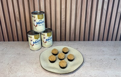 Prato com docinhos cobertos com farofa crocante e latas de Leite Moça empilhadas ao fundo.