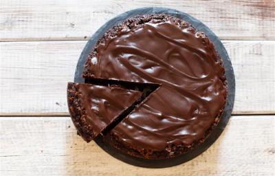 Fotografia em tons de marrom e branco de uma bancada marrom vista de cima, contém um prato redondo e branco com um bolo de chocolate com cobertura de chocolate e uma fatia sendo retirada por uma espátula Fotografia em tons de marrom e branco de uma bancada marrom vista de cima, contém um prato redondo e branco com um bolo de chocolate com cobertura de chocolate e uma fatia sendo retirada por uma espátula