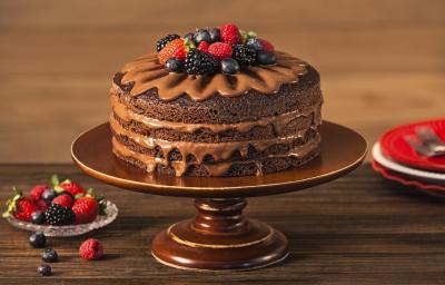 Fotografia em tons de marrom em uma bancada de madeira escura com um suporte para bolo marrom e o naked cake com ganache de cacau e frutas vermelhas em cima. Ao lado, um pratinho de vidro com frutas vermelhas.