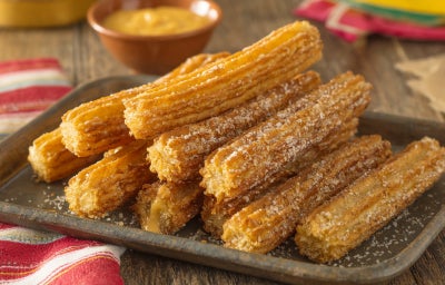 imagem mostra churros dourados empanados em açúcar e canela, servidos com doce de leite.