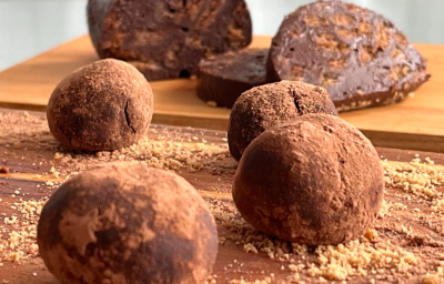 A imagem mostra quatro bolinhas de chocolate em formato de trufa sobre uma tábua de madeira. Ao fundo, uma massa de chocolate em formato cilindrico está cortada em fatias.
