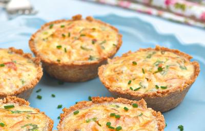 Fotografia em tons de azul em uma mesa de madeira azul com um prato redondo raso azul e alguns mini quiches de batata e salmão.