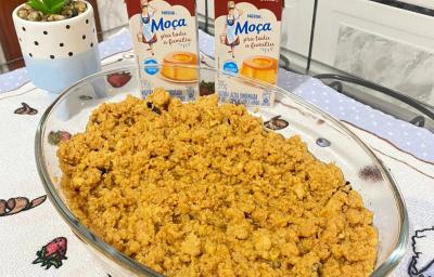 Foto da receita de ambrosia em tom amarelo, numa travessa de vidro, sobre uma mesa com toalha e nela há duas caixas do produto de Moça e uma vasinho com uma planta em tom verde Foto da receita de ambrosia em tom amarelo, numa travessa de vidro, sobre uma mesa com toalha e nela há duas caixas do produto de Moça e uma vasinho com uma planta em tom verde