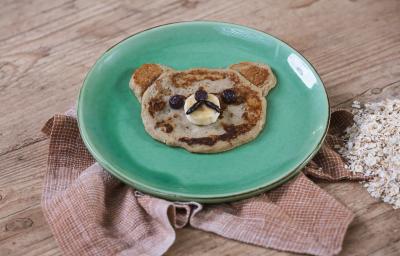 Foto da receita de Panqueca de Banana. Observa-se um pratinho verde e a panquequinha com cara de ursinho. Foto da receita de Panqueca de Banana. Observa-se um pratinho verde e a panquequinha com cara de ursinho.