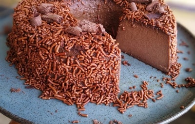 Fotografia de um prato raso, grande, de vidro azul suspenso com o brigadeirão de café sobre ele. O brigadeirão está com um pedaço grande cortado, mostrando a textura da sobremesa, que está finalizada com granulado e raspas de chocolate. Fotografia de um prato raso, grande, de vidro azul suspenso com o brigadeirão de café sobre ele. O brigadeirão está com um pedaço grande cortado, mostrando a textura da sobremesa, que está finalizada com granulado e raspas de chocolate.