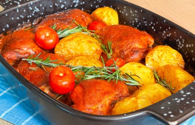 Fotografia mostra uma assadeira com sobrecoxas assadas com alecrim, batatas douradas e tomates cereja.