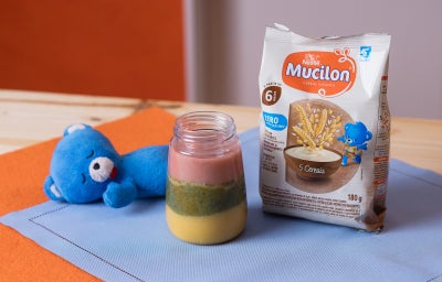 Fotografia em tons de azul e laranja em uma bancada de madeira. Ao centro, um copo de vidro com uma bebida colorida, rosa, amarela e verde.