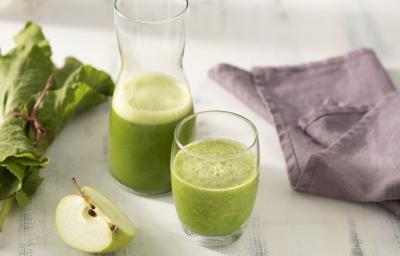 Fotografia em tons de verde e roxo de uma bancada branca, sobre ela dois copos de vidro com suco verde. Ao lado um paninho roxo, uma fatia de maçã verde e folhas de couve. Fotografia em tons de verde e roxo de uma bancada branca, sobre ela dois copos de vidro com suco verde. Ao lado um paninho roxo, uma fatia de maçã verde e folhas de couve.