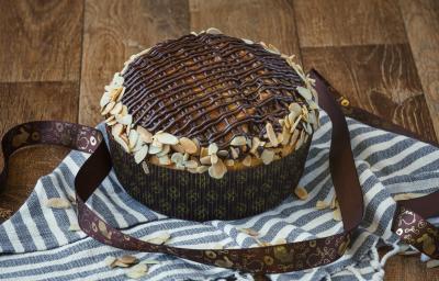 Imagem da receita de Colomba Pascal, decorada com tiras de chocolate e amêndoas em lascas, sobre uma bancada decorada com um tecido azul e branco, com uma fita de páscoa decorativa Imagem da receita de Colomba Pascal, decorada com tiras de chocolate e amêndoas em lascas, sobre uma bancada decorada com um tecido azul e branco, com uma fita de páscoa decorativa