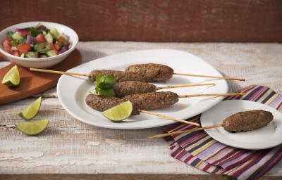 fotografia em tons de bege, marrom e branco de uma bancada bege vista de frente, contém um prato branco redondo com palitos de bolinhos de carne moída e fatias de limão, ao lado um pano com um prato branco e redondo com um espetinho com a kafta