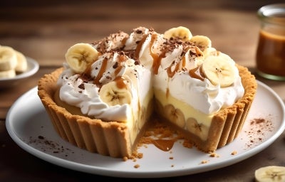 Em cima de uma mesa, prato com fatia de banoffee com massa de biscoitos crocante, bananas em rodelas, doce de leite, decorado com chantilly cremoso, banana em rodelas e cacau em pó. Em cima de uma mesa, prato com fatia de banoffee com massa de biscoitos crocante, bananas em rodelas, doce de leite, decorado com chantilly cremoso, banana em rodelas e cacau em pó.