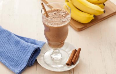 Fotografia em tons de marrom em uma bancada de madeira clara com um pano azul ao lado e ao centro um pratinho branco com uma taça de vidro e o smoothie de cacau e banana dentro. Ao fundo, um cacho de banana apoiado em uma tábua de madeira.