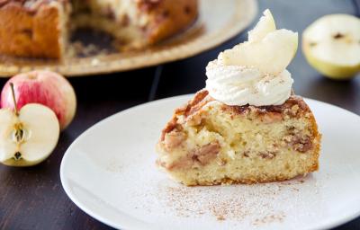Fotografia em tons de branco em uma bancada de madeira de cor preta. Ao centro, um prato branco contendo a torta e ao fundo, uma maçã cortada ao meio Fotografia em tons de branco em uma bancada de madeira de cor preta. Ao centro, um prato branco contendo a torta e ao fundo, uma maçã cortada ao meio