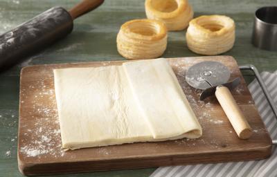 Fotografia em tons de marrom em uma bancada de madeira verde, uma tábua de madeira suja com farinha e um pedaço de massa folhada e um cortador de pizza ao lado. Fotografia em tons de marrom em uma bancada de madeira verde, uma tábua de madeira suja com farinha e um pedaço de massa folhada e um cortador de pizza ao lado.