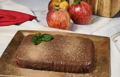 Fotografia em tons de branco e vermelho em uma bancada de mármore e um prato retangular e marrom ao centro. Em cima dele existe uma torta mousse de chocolate polvilhada com cacau em pó. Ao fundo existe 3 maçãs. Fotografia em tons de branco e vermelho em uma bancada de mármore e um prato retangular e marrom ao centro. Em cima dele existe uma torta mousse de chocolate polvilhada com cacau em pó. Ao fundo existe 3 maçãs.