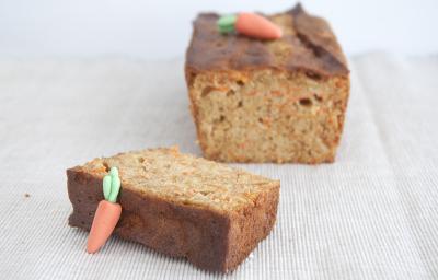Fotografia em tons claros de um bolo de cenoura com mini cenouras em cima, feitas de pasta americana, cortado em uma fatia grossa que está mais no centro da foto. O bolo está sobre uma toalha de mesa na cor marrom claro. Fotografia em tons claros de um bolo de cenoura com mini cenouras em cima, feitas de pasta americana, cortado em uma fatia grossa que está mais no centro da foto. O bolo está sobre uma toalha de mesa na cor marrom claro.