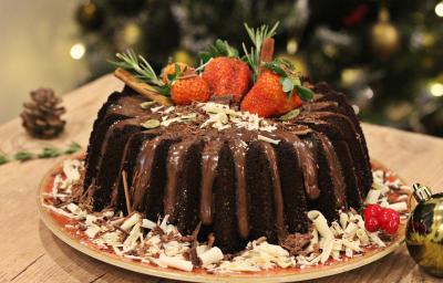 Fotografia em tons de marrom e vermelho de uma bancada de madeira clara, sobre ela um prato vermelho redondo com o bolo de chocolate decorado. Ao lado uma bolinha natalina e uma pinha. Fotografia em tons de marrom e vermelho de uma bancada de madeira clara, sobre ela um prato vermelho redondo com o bolo de chocolate decorado. Ao lado uma bolinha natalina e uma pinha.