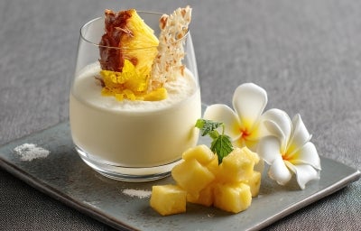 Imagem de um copo servido com um creme cremoso de abacaxi, decorado com uma fatia de abacaxi em meia-lua e lascas de coco.