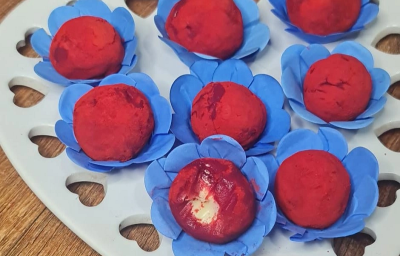 Foto em tons de vermelho da receita de brigadeiro red velvet servida em diversas porções sobre embalagens azuis, que se dispõem em cima de uma base em formato de coração branca sobre uma mesa de madeira