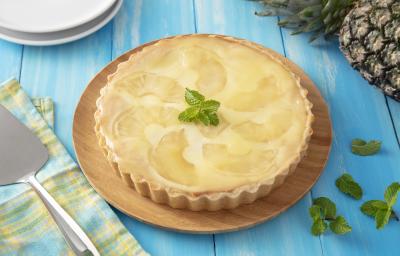 Receita de Torta de Abacaxi ao Creme de Ricota. Observa-se uma torta inteira redonda decorada com hortelã e abacaxi sobre uma tábua redonda de madeira em uma superfície de madeira azul clara. Ao lado esquerdo, uma espátula prateada. Receita de Torta de Abacaxi ao Creme de Ricota. Observa-se uma torta inteira redonda decorada com hortelã e abacaxi sobre uma tábua redonda de madeira em uma superfície de madeira azul clara. Ao lado esquerdo, uma espátula prateada.