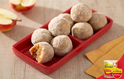 Doces de maçã em formato de bolinhas, cobertos com açúcar, dispostos em travessa vermelha, com maçãs ao fundo.