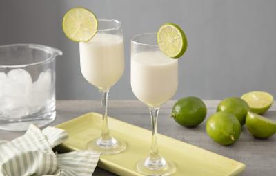 Foto em tons claros de verde da receita de nevada com leite MOÇA servida em duas taças largas e altas sobre uma base de porcelana verde, ao lado há um pano listrado nos tons da foto e ao fundo há limões e um ricipiente de vidro com gelo. Foto em tons claros de verde da receita de nevada com leite MOÇA servida em duas taças largas e altas sobre uma base de porcelana verde, ao lado há um pano listrado nos tons da foto e ao fundo há limões e um ricipiente de vidro com gelo.