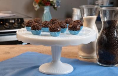 Fotografia de dez forminhas azuis com brigadeiros passados no granulado e apoiados em um enfeite para bolo branco e alto. Ao fundo tem um fogão, um vaso azul com flores, e ao lado do brigadeiro tem duas jarras decorativas de vidro com açúcar e cacau. Fotografia de dez forminhas azuis com brigadeiros passados no granulado e apoiados em um enfeite para bolo branco e alto. Ao fundo tem um fogão, um vaso azul com flores, e ao lado do brigadeiro tem duas jarras decorativas de vidro com açúcar e cacau.