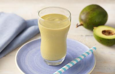 Fotografia em tons de azul e verde de uma bancada com um pratinho azul, um copo transparente com uma vitamina de abacate e um canudo. Ao fundo um paninho azul e um abacate. Fotografia em tons de azul e verde de uma bancada com um pratinho azul, um copo transparente com uma vitamina de abacate e um canudo. Ao fundo um paninho azul e um abacate.