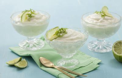 Fotografia em tons de azul e verde de uma bancada azul claro, com duas fatias de limão, um pano verde claro dobrado, duas colheres douradas, um limão pela metade e três taças transparentes com a mousse e raspas e rodelas de limão para decorar.