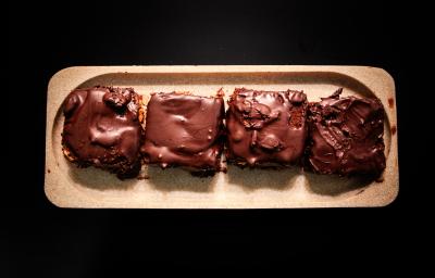 Fotografia vista de cima de quatro porções unidades de pão de mel com ganache de pitanga e cobertura de chocolate amargo. A sobremesa está em um apoio de madeira decorativo retangular, o qual está sobre uma mesa preta.