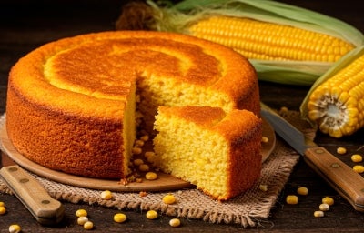 Fotografia de um bolo de milho dourado, com uma fatia cortada, revelando seu interior macio. Está sobre uma tábua de madeira, cercado por grãos de milho e espigas frescas.