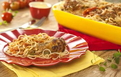 Fotografia em tons de amarelo e vermelho em uma bancada de madeira clara, panos amarelo e vermelho, um recipiente amarelo com o macarrão espaguete com asa de frango dentro dele, um prato vermelho com um pouco do macarrão.