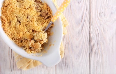 Fotografia em tons claros visto de cima de uma torta cremosa de farofa doce sobre um pano amarelo e branco dobrado, apoiado em uma mesa de madeira clara. Fotografia em tons claros visto de cima de uma torta cremosa de farofa doce sobre um pano amarelo e branco dobrado, apoiado em uma mesa de madeira clara.