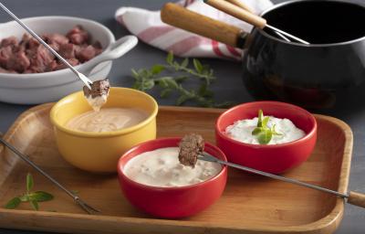 Fotografia em tons de vermelho e amarelo de uma bancada. A frente 1 pote amarelo com molho e 2 potes vermelhos com molho, 2 espetos com pedaços de carne na ponta e ao fundo uma travessa com pedaços de carne crua e uma panela de fondue preta. Fotografia em tons de vermelho e amarelo de uma bancada. A frente 1 pote amarelo com molho e 2 potes vermelhos com molho, 2 espetos com pedaços de carne na ponta e ao fundo uma travessa com pedaços de carne crua e uma panela de fondue preta.