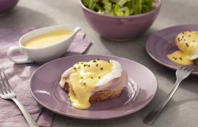 Fotografia em tons de lilás em uma bancada de madeira clara, um paninho lilás, um prato redondo lilás com o ovo benedict em cima dele, com a fatia de pão tostado, presunto, o ovo e cebolinha-verde para decorar. Ao fundo, um bowl com salada verde. Fotografia em tons de lilás em uma bancada de madeira clara, um paninho lilás, um prato redondo lilás com o ovo benedict em cima dele, com a fatia de pão tostado, presunto, o ovo e cebolinha-verde para decorar. Ao fundo, um bowl com salada verde.