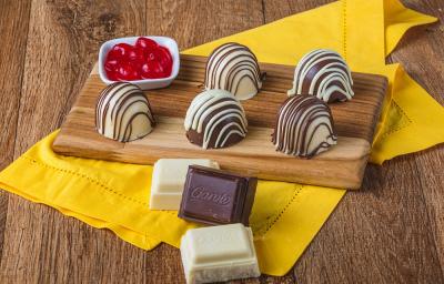 Fotografia em tons de marrom em uma bancada de madeira de cor marrom. Ao centro, um pano amarelo contendo uma tábua de madeira com as trufas e ao lado, há um recipiente contendo cerejas. Fotografia em tons de marrom em uma bancada de madeira de cor marrom. Ao centro, um pano amarelo contendo uma tábua de madeira com as trufas e ao lado, há um recipiente contendo cerejas.