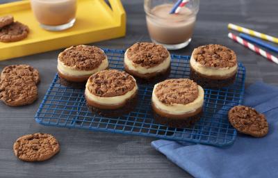 Foto de uma bancada cinza com uma grade contendo cinco doces circulares em camadas diferentes e decorados com cookies Nescau. Além disso há um tecido azul à direita e, ao fundo, canudos coloridos de papel, uma bandeja amarela e copos com leite e Nescau