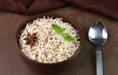 Fotografia em tons escuros de um recipiente fundo na cor marrom com arroz e especiarias e duas folhas de hortelã por cima. Ao lado, uma colher de sopa desenhada, sobre uma mesa marrom em tom escuro. Fotografia em tons escuros de um recipiente fundo na cor marrom com arroz e especiarias e duas folhas de hortelã por cima. Ao lado, uma colher de sopa desenhada, sobre uma mesa marrom em tom escuro.