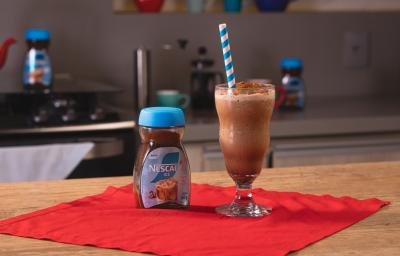 Fotografia de um copo de vidro grande com frozen de café, ao lado de uma embalagem de nescafé gelado, sobre um pano vermelho e uma mesa de madeira. Dentro do copo tem um canudo azul e branco apoiado. Fotografia de um copo de vidro grande com frozen de café, ao lado de uma embalagem de nescafé gelado, sobre um pano vermelho e uma mesa de madeira. Dentro do copo tem um canudo azul e branco apoiado.