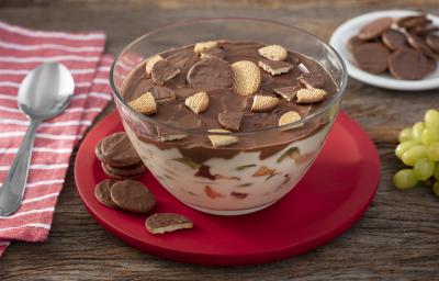 Foto da receita de Taça Cremosa de Calipso, feita em uma taça de vidro grande, sobre um prato vermelho com alguns biscoitos ao redor, sobre uma bancada de madeira decora com um guardanapo de tecido e com algumas uvas verdes ao lado
