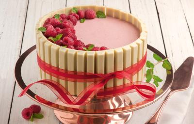 Fotografia em tons de rosa em uma bancada de madeira clara, um suporte para bolo em vidro rosado, com a mousse de framboesa com KitKat Branco ao redor, formando uma linda torta. Fotografia em tons de rosa em uma bancada de madeira clara, um suporte para bolo em vidro rosado, com a mousse de framboesa com KitKat Branco ao redor, formando uma linda torta.