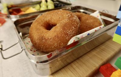 Fotografia em tons de vermelho em uma bancada de madeira clara, uma marmita ao meio com dois donuts de maçã dentro. Ao lado, brinquedos infantis. Fotografia em tons de vermelho em uma bancada de madeira clara, uma marmita ao meio com dois donuts de maçã dentro. Ao lado, brinquedos infantis.