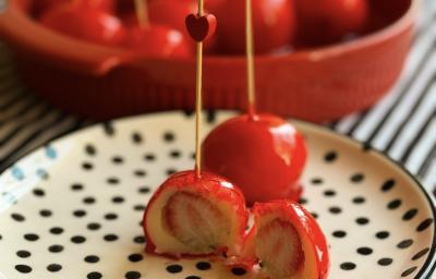 Fotografia de dois morangos do amor prontos em um prato raso branco com bolinhas pretas. Um dos morangos está cortado na metade, mostrando o interior. Ao fundo tem mais porções dentro de um recipiente grande vermelho. Fotografia de dois morangos do amor prontos em um prato raso branco com bolinhas pretas. Um dos morangos está cortado na metade, mostrando o interior. Ao fundo tem mais porções dentro de um recipiente grande vermelho.