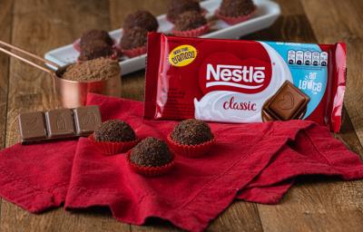 Foto aproximada da receita de brigadeiro gourmet, em uma bancada de madeira decorada com um tecido vermelho, uma barra de chocolate ao leite e mais brigadeiros sobre um prato branco