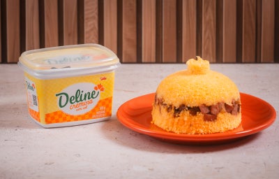 Foto de um cuscuz recheado com charque, servido em um prato laranja, ao lado de uma embalagem de margarina deline